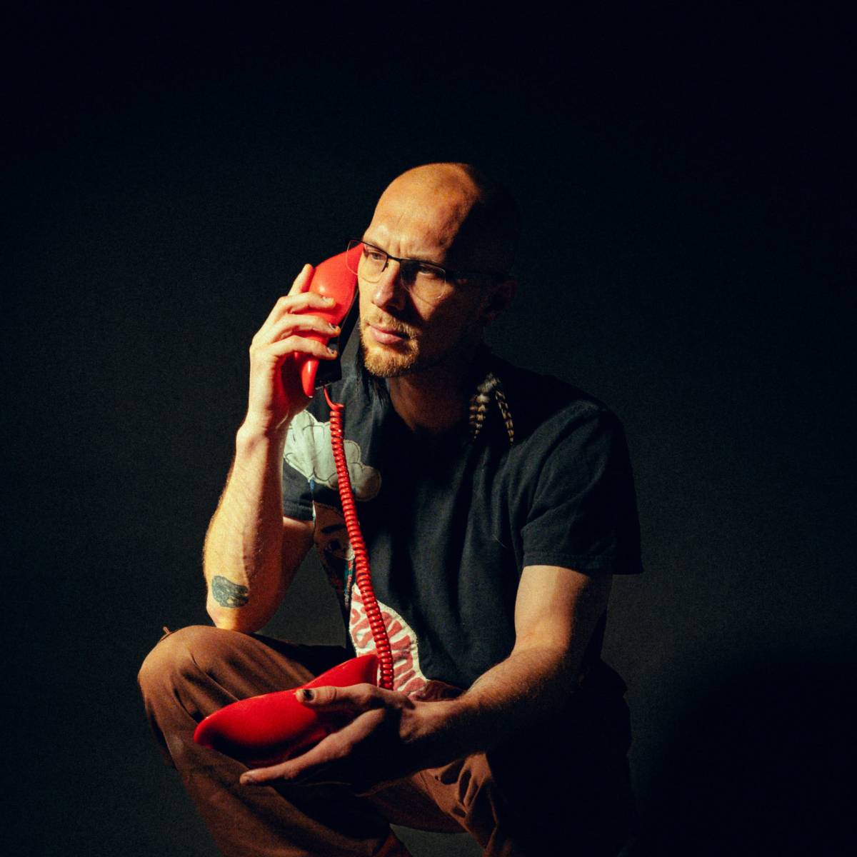 A man with glasses is squatting on a wooden stool while talking on a red telephone.