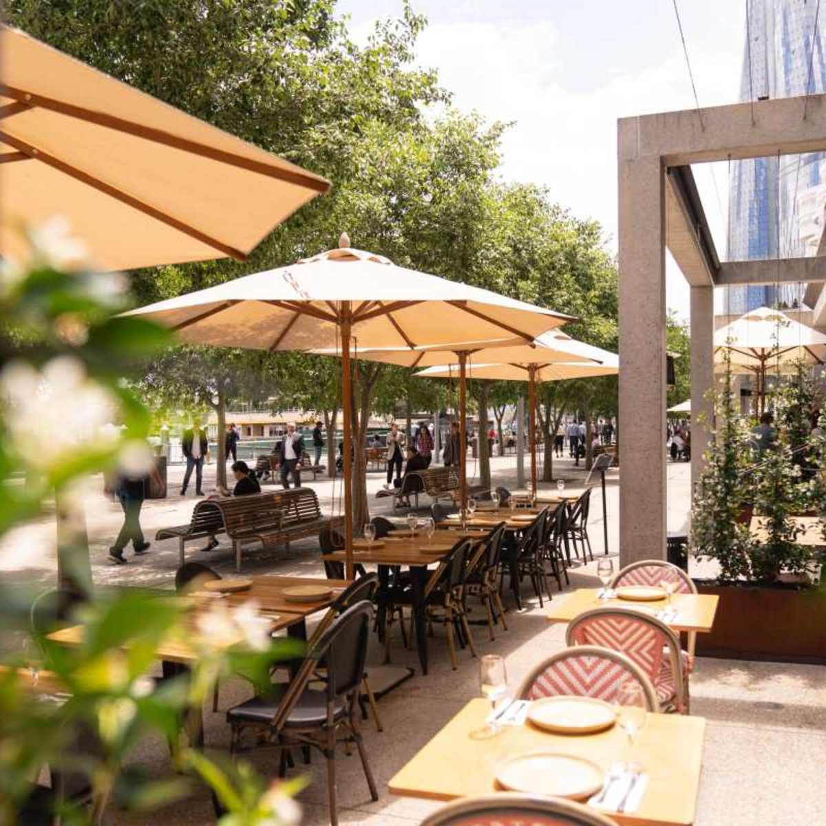 A street-side restaurant patio features wooden tables and umbrellas, surrounded by greenery and pedestrians.