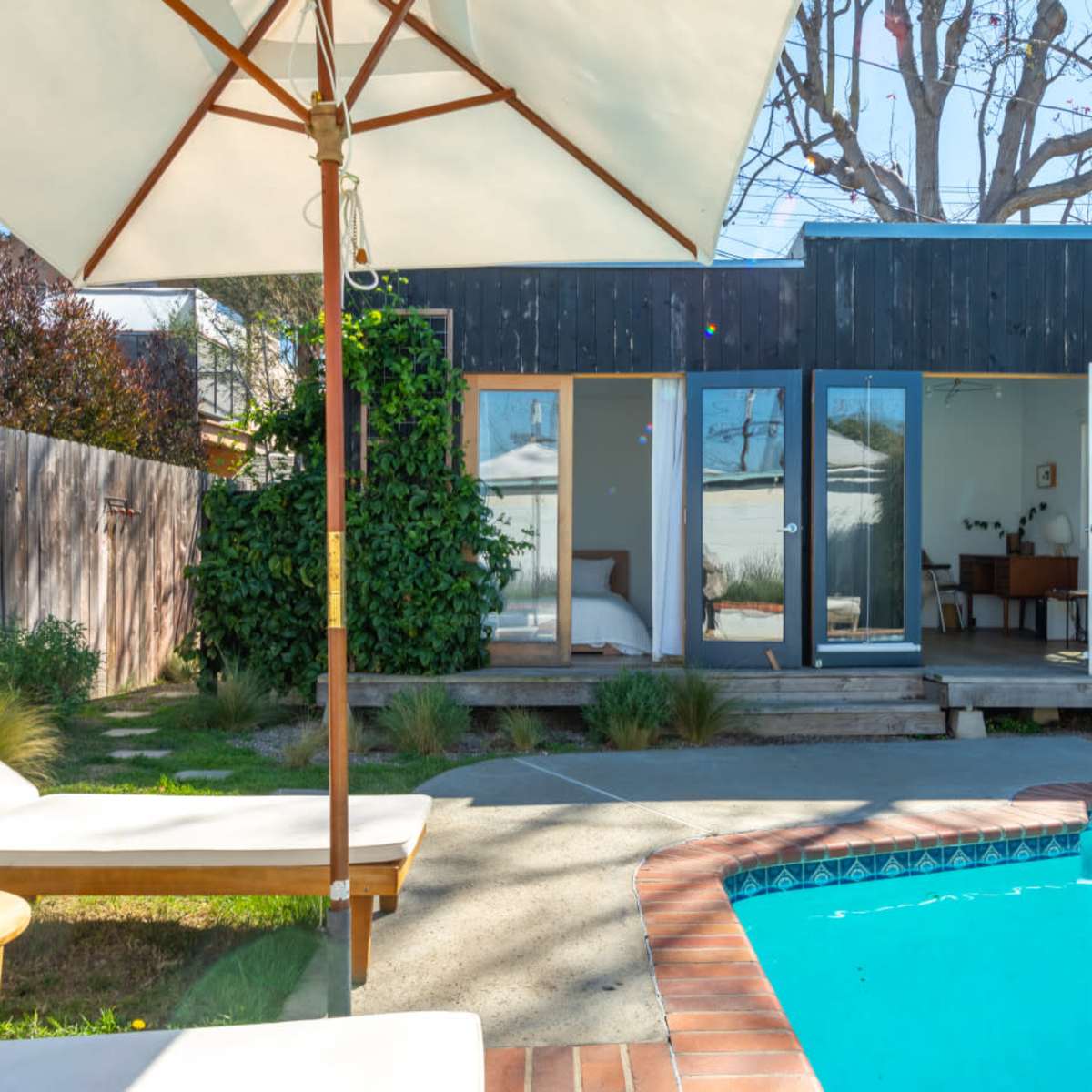 A small, modern cabin with large glass doors overlooks a landscaped yard and a swimming pool, surrounded by a wooden fence and trees.