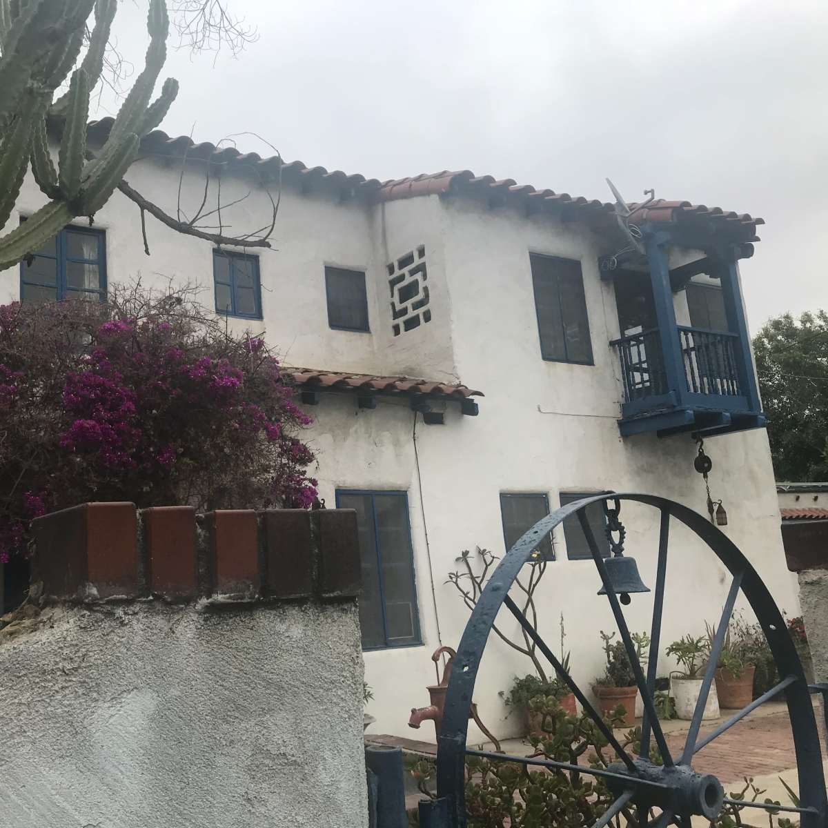 A two-story building with blue railings and a stucco exterior is surrounded by a cactus and flowering plants.