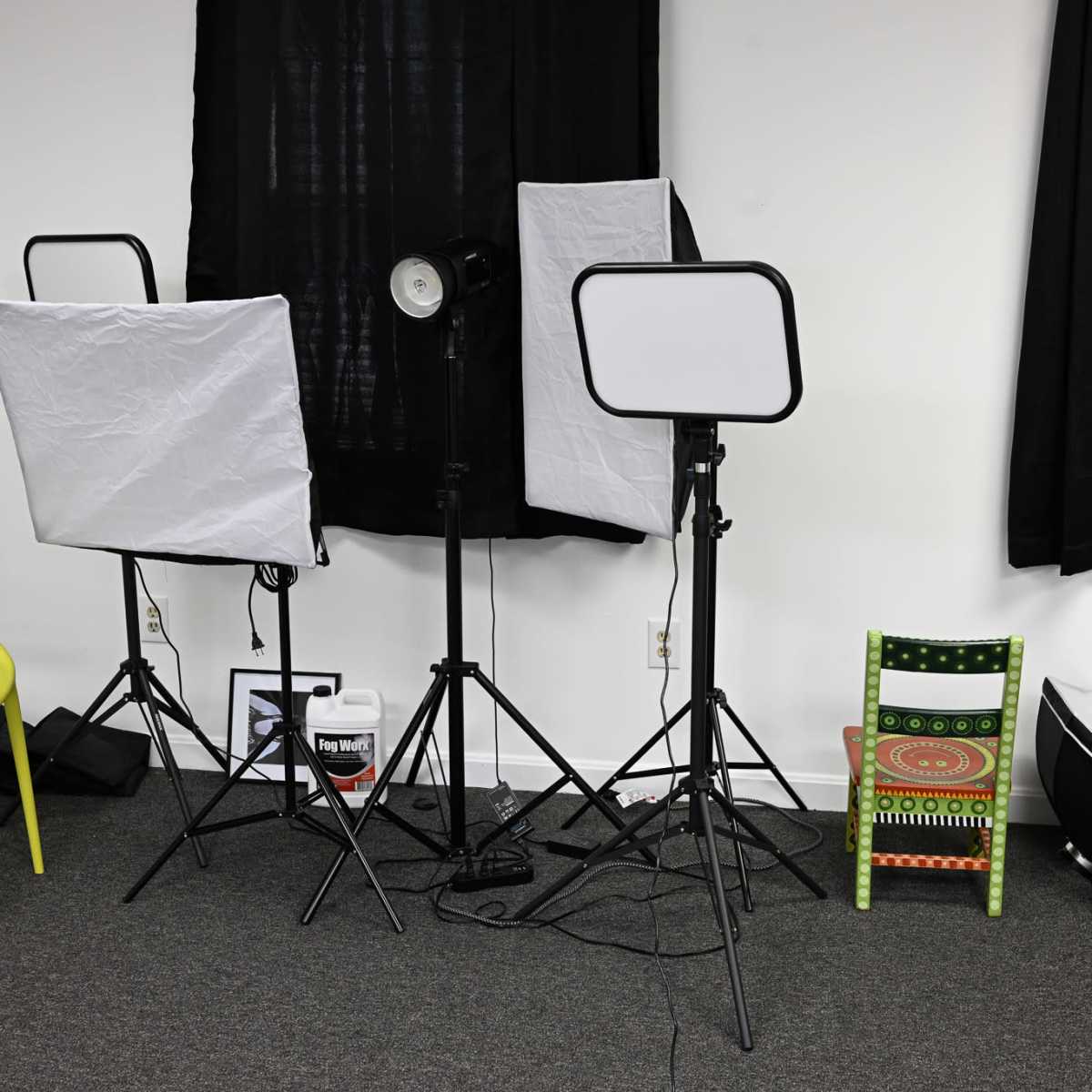 A photography setup with two softbox light stands, a spotlight, and colorful chairs in a room with black curtains.