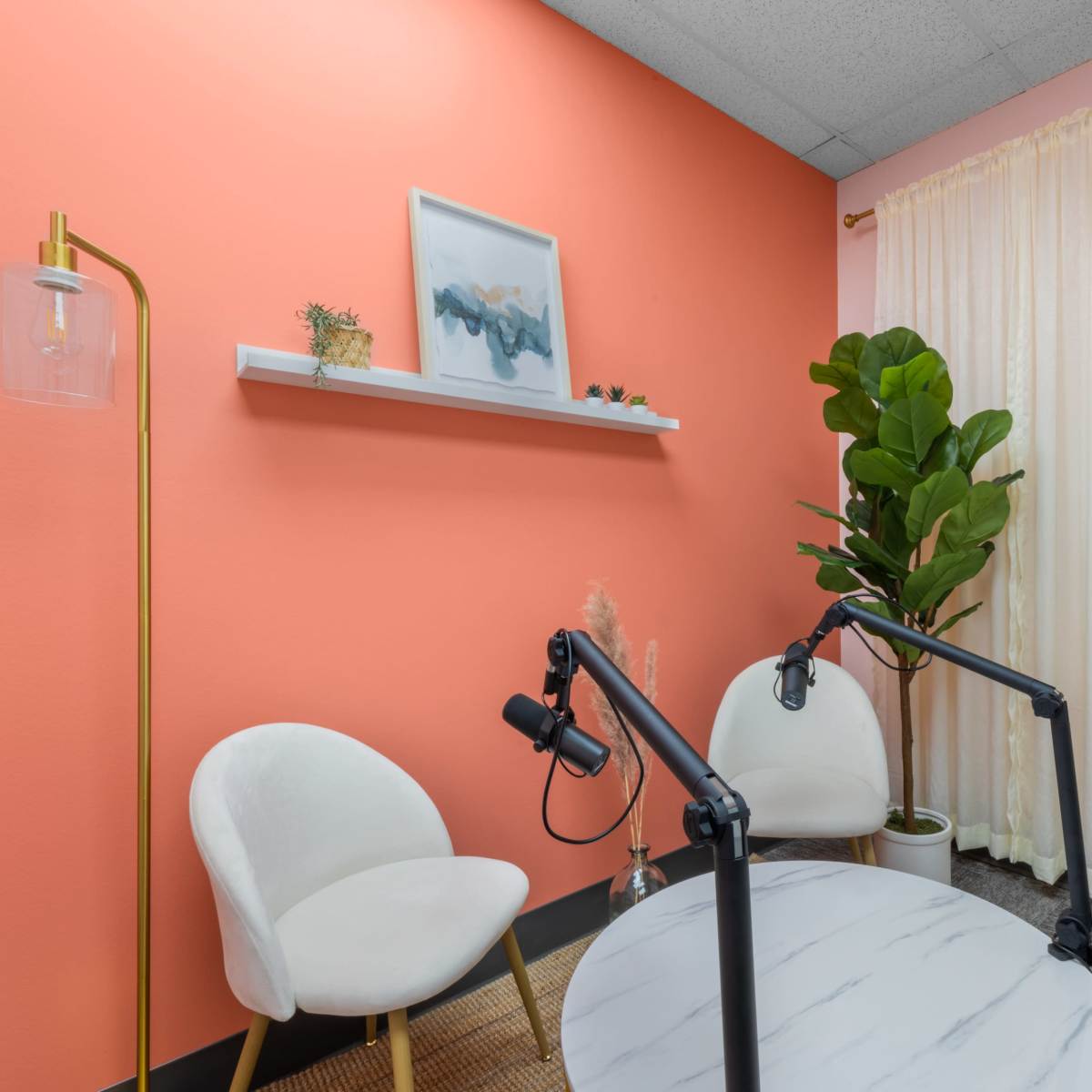 A small room with coral-colored walls, two white chairs, a marble-top table, and a potted plant beside a window with sheer curtains.