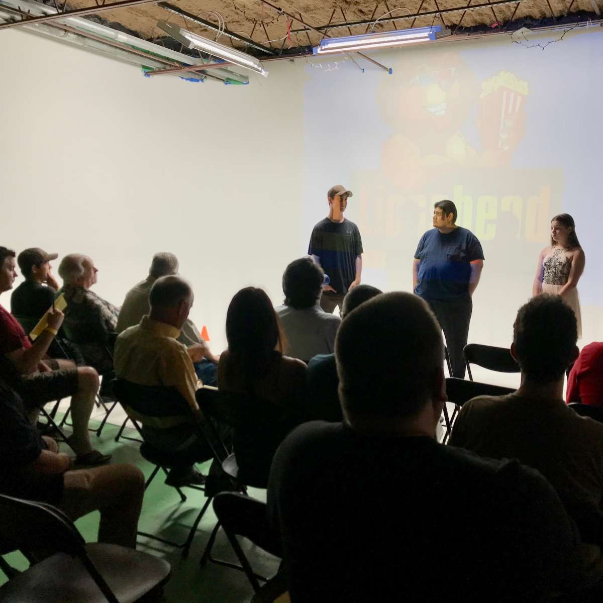A group of people sits in chairs while three individuals stand in front of a large projector screen displaying an animated character and text.
