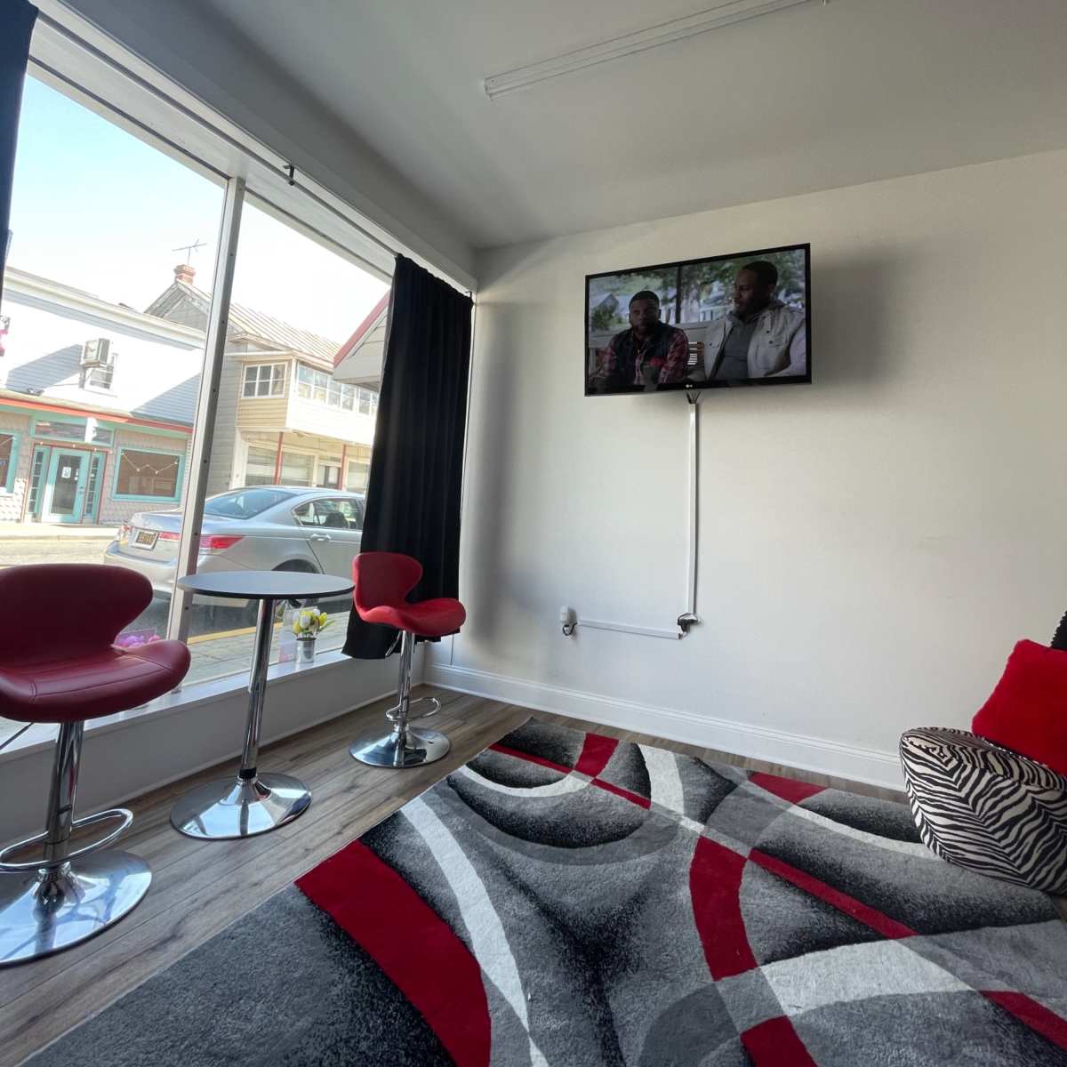 A modern indoor space with two red bar stools in front of a television mounted on the wall, beside a large window and a patterned rug on the floor.