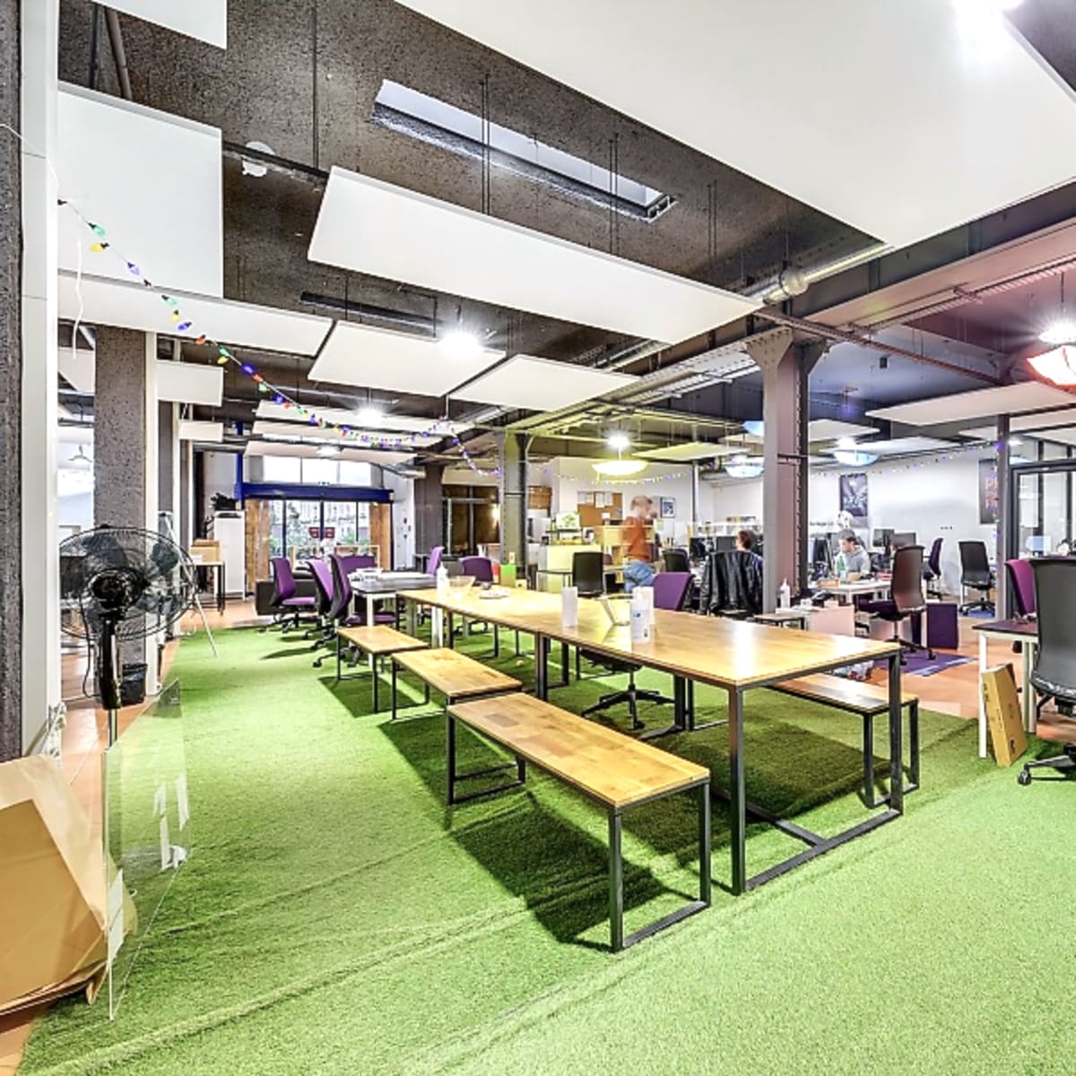 An open office space with long tables, chairs, and computer workstations, and artificial grass covering the floor.