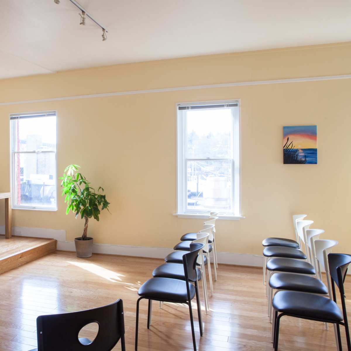 A bright, empty room featuring rows of chairs facing a small stage with a wooden platform, a bookshelf filled with books, a television mounted on the wall, and large windows letting in natural light.