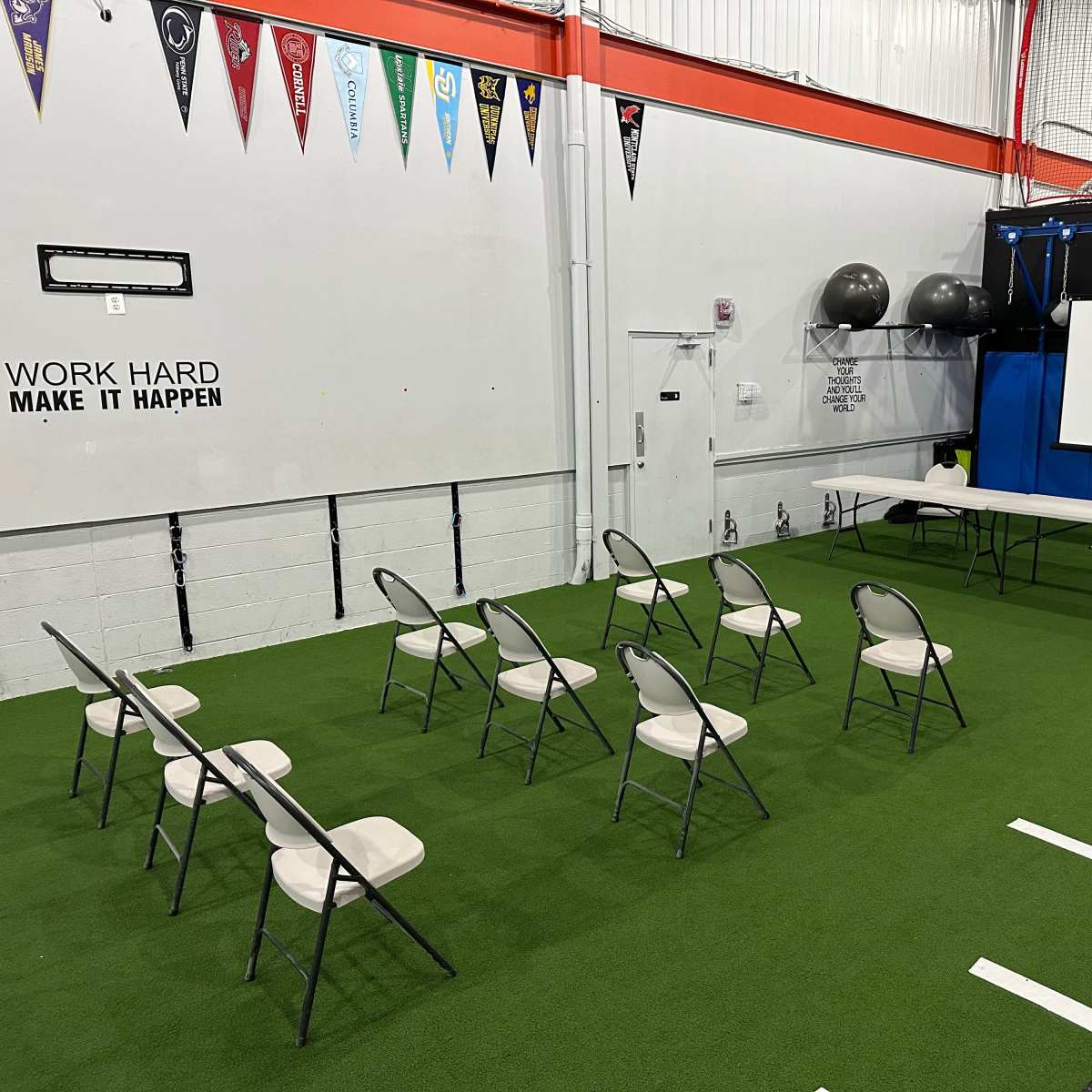 A gymnasium setup features rows of chairs facing a long table and a large screen, with exercise equipment in the background.