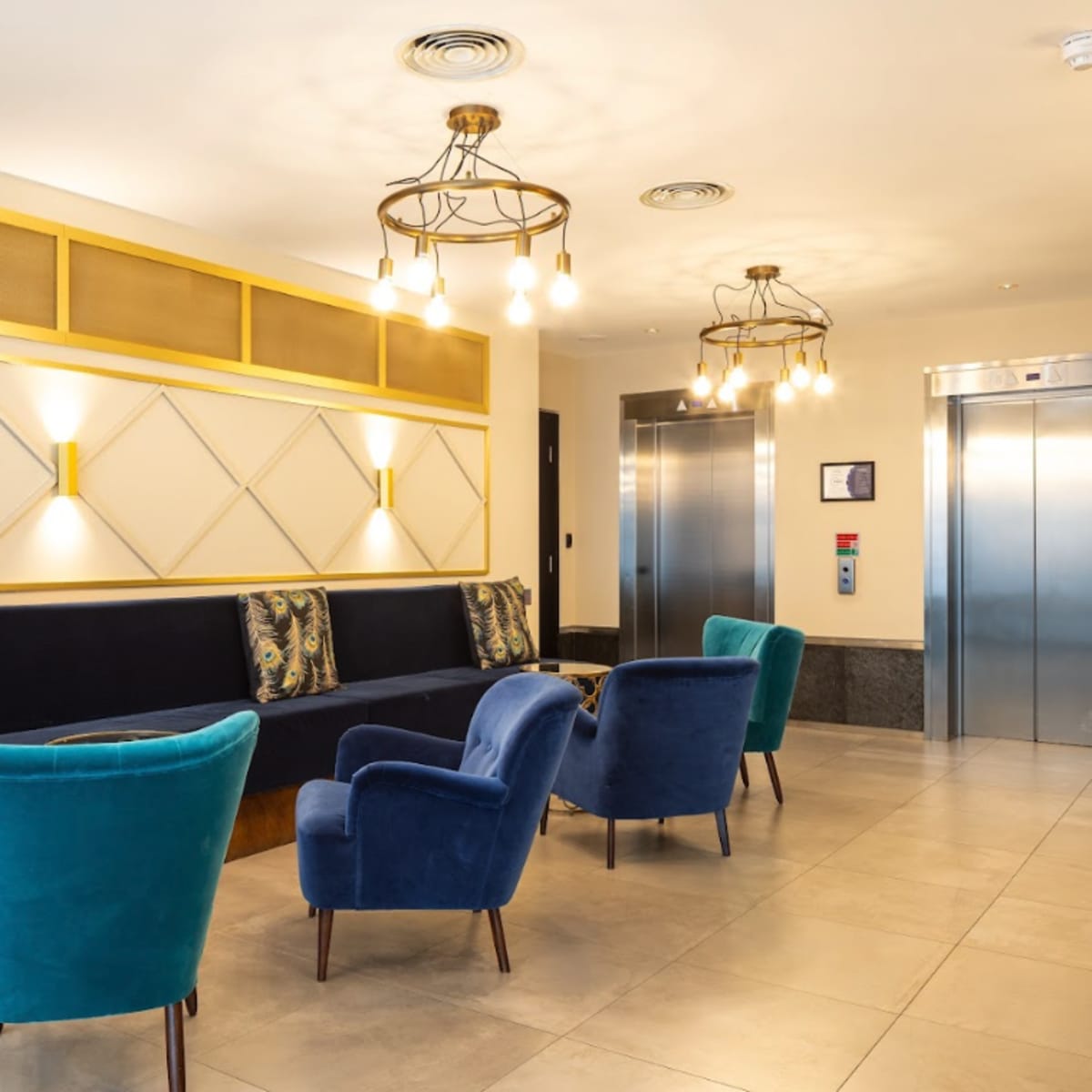 A modern lobby area with a seating arrangement featuring blue and teal chairs, decorative cushions, and two elevators in the background.