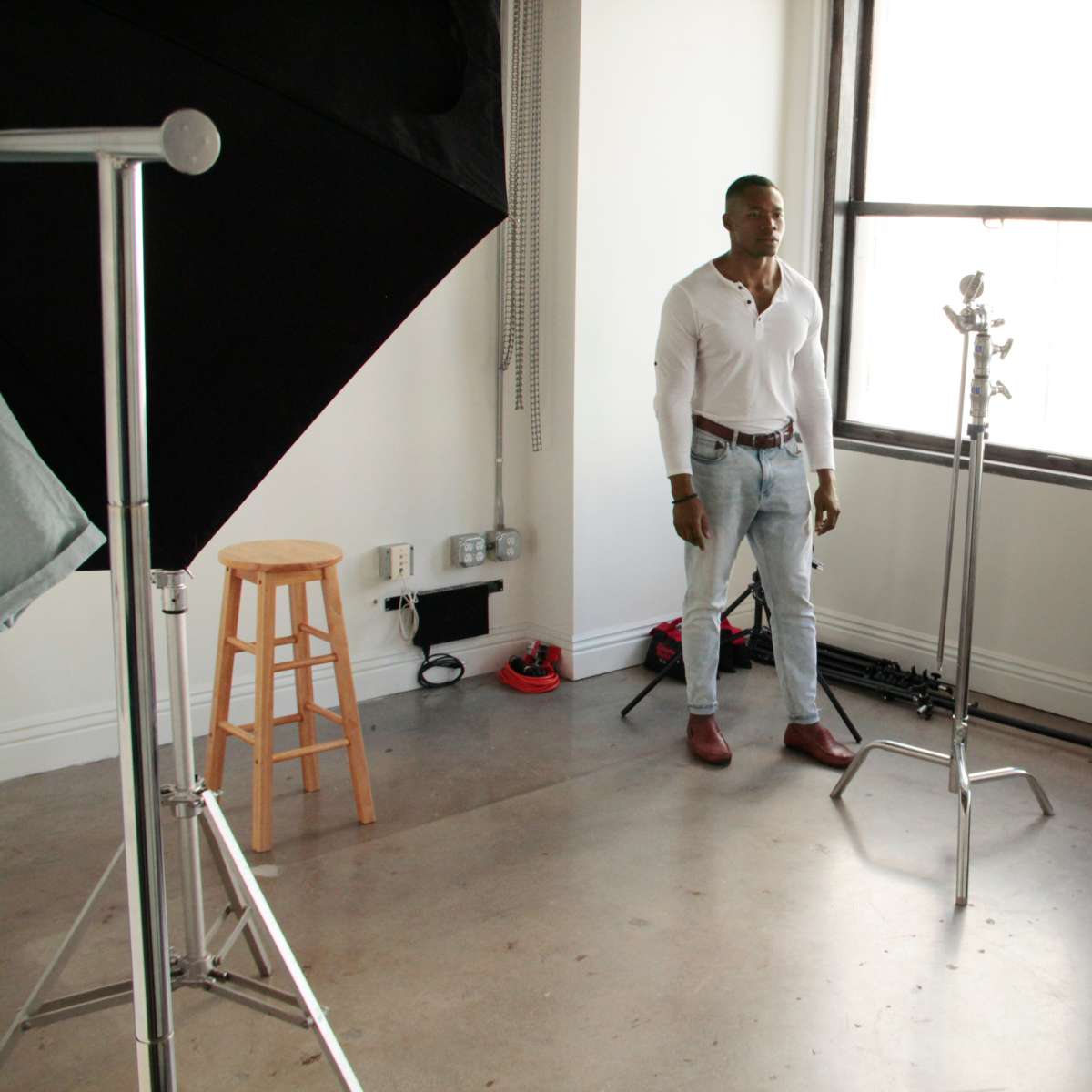 A man stands in a photography studio filled with equipment, including a stool, light stands, and a reflector.