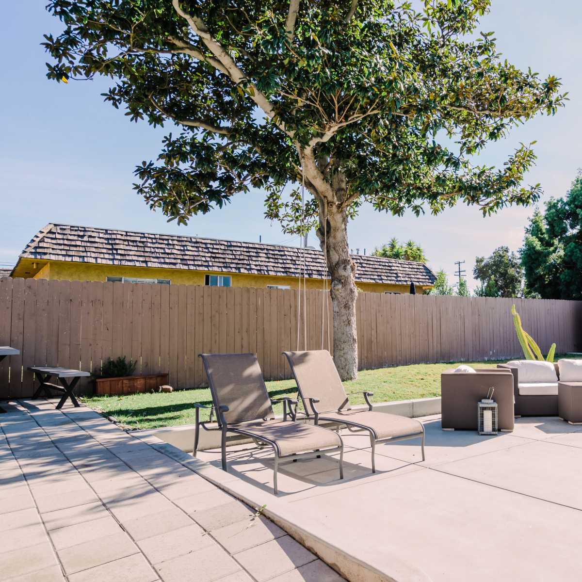 A backyard setting with patio furniture arranged on a concrete surface, surrounded by a wooden fence and a tree.