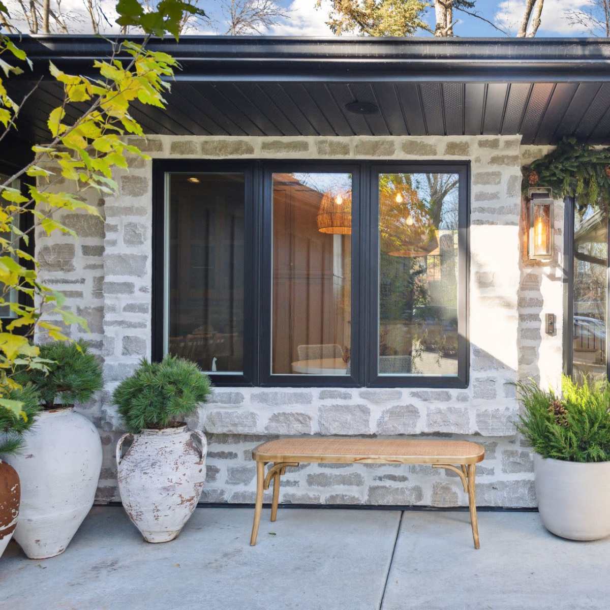 A front porch with a stone wall, large windows, a decorated door with a green wreath, a wooden bench, and potted plants.