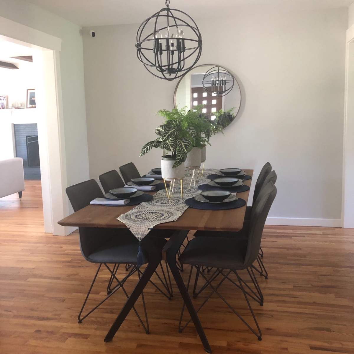 A wooden dining table with eight black chairs is set with blue plates and a decorative runner, beneath a round chandelier and a circular mirror on the wall.