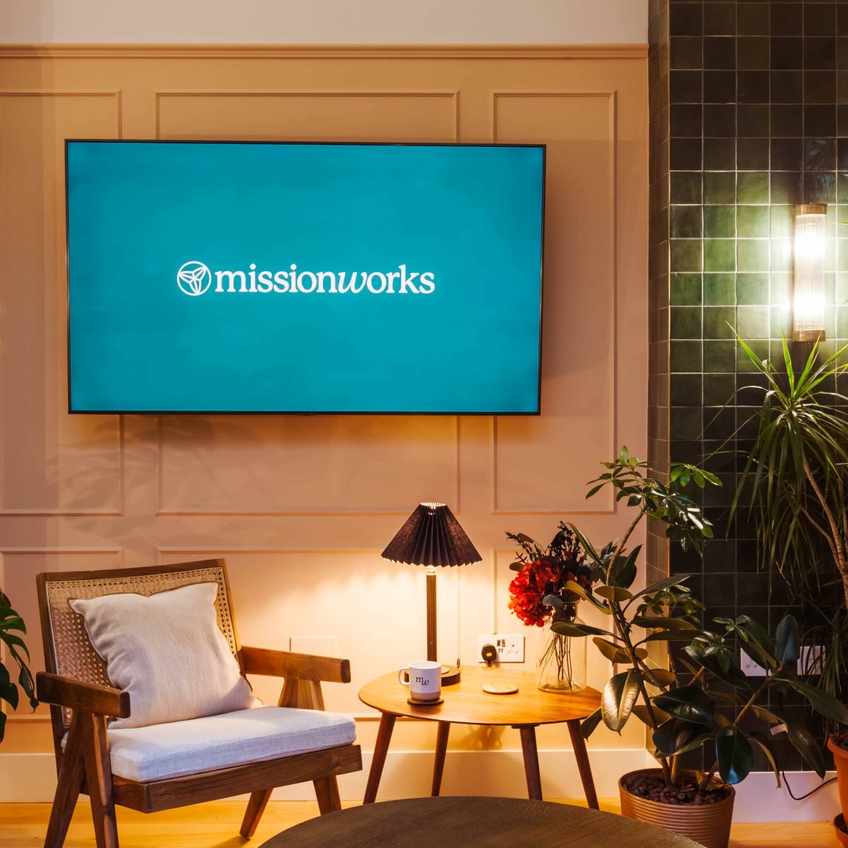 A modern lounge area features a large television displaying the "missionworks" logo above a wooden chair and a side table, surrounded by potted plants.