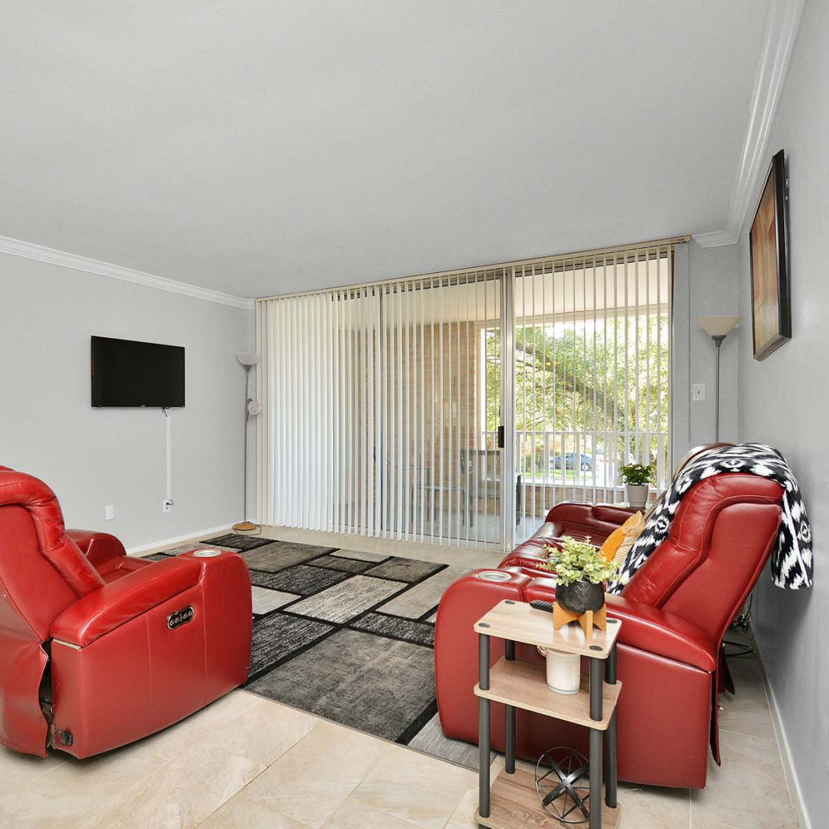 A living room with two red recliners facing a small table, a flat-screen TV mounted on the wall, and sliding glass doors leading to a balcony.