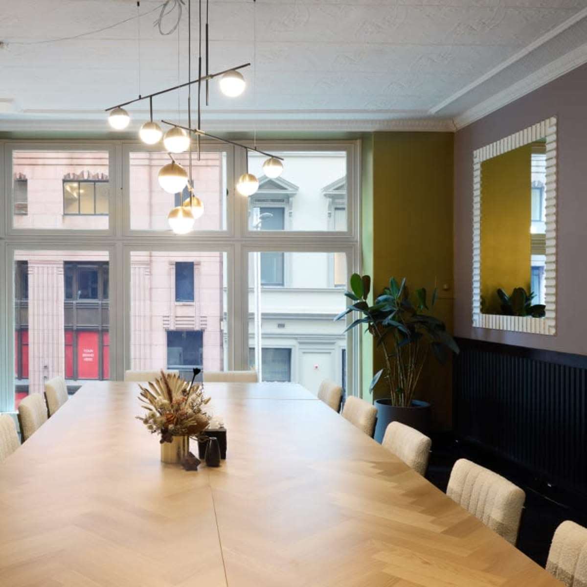 A long wooden conference table is positioned in a brightly lit room with large windows, potted plants, and modern lighting fixtures.
