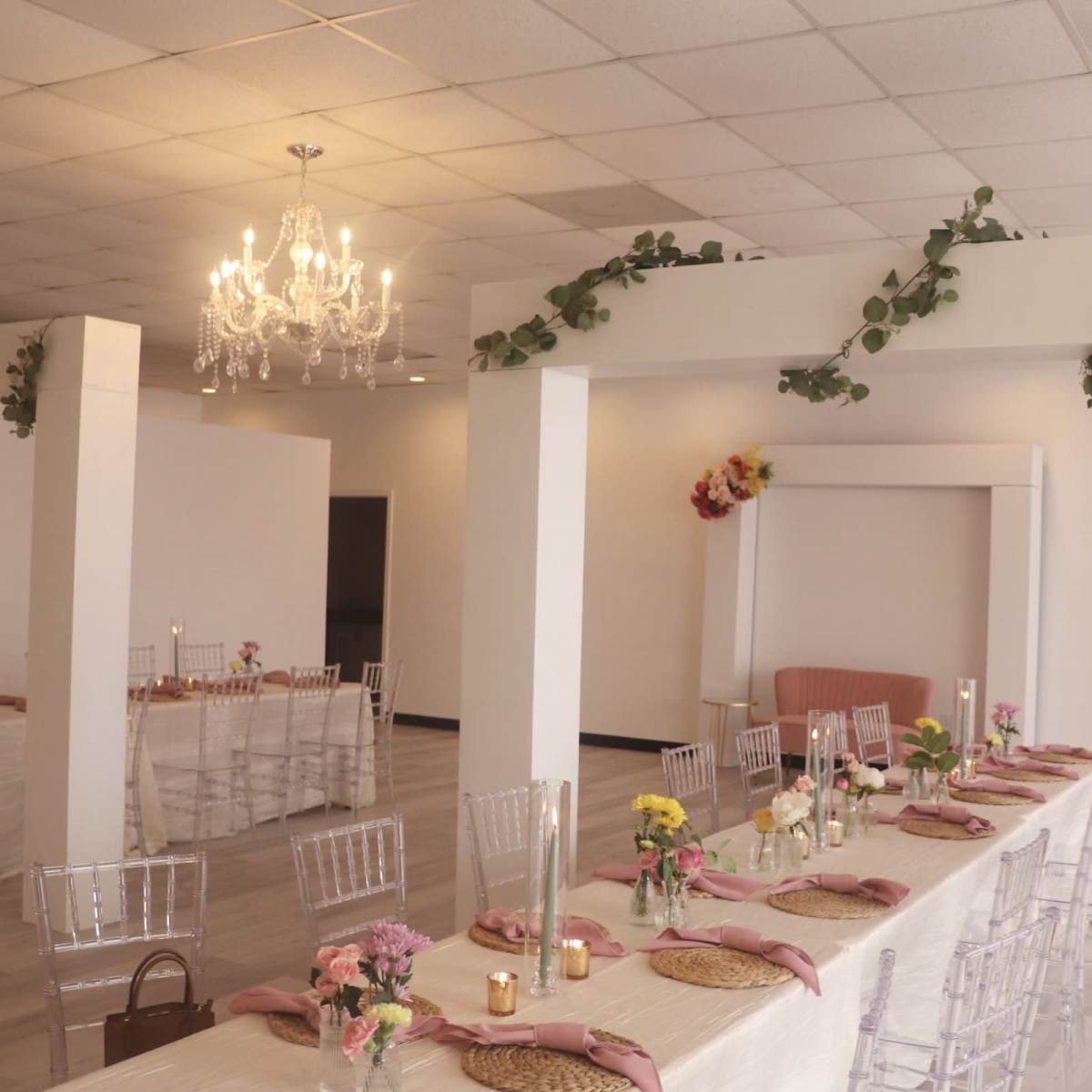 A spacious, elegantly decorated event venue features long tables adorned with floral centerpieces and soft lighting under a chandelier.
