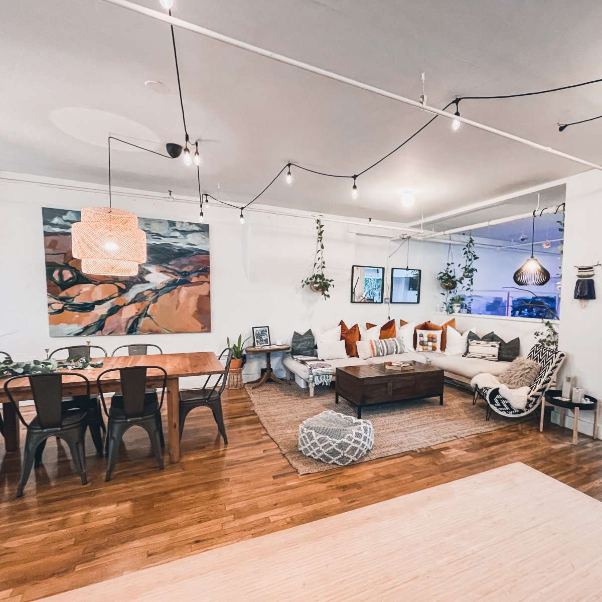 A spacious, modern living area with a large sectional sofa, a wooden dining table, and decorative plants hanging from the ceiling.