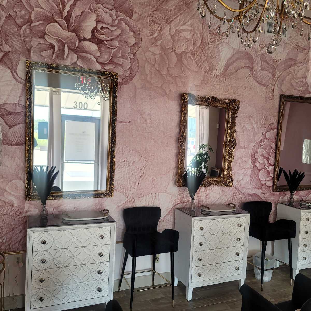A stylish interior featuring three ornate mirrors above white dressers, flanked by black chairs and a lamp on a gold stand, all against a floral-patterned pink wallpaper.