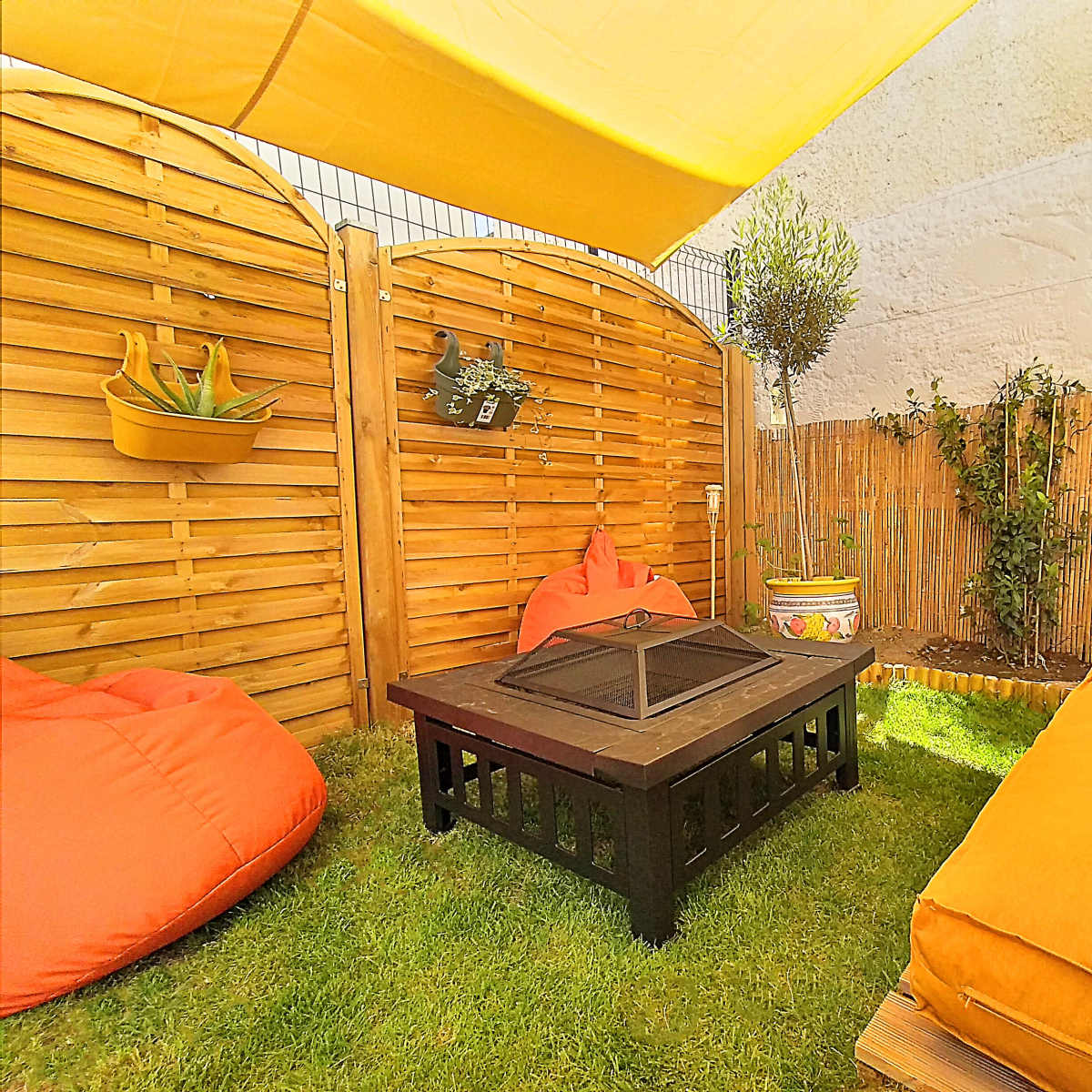 A cozy outdoor seating area with a black coffee table, bright orange cushions, and wooden privacy screens surrounded by greenery.