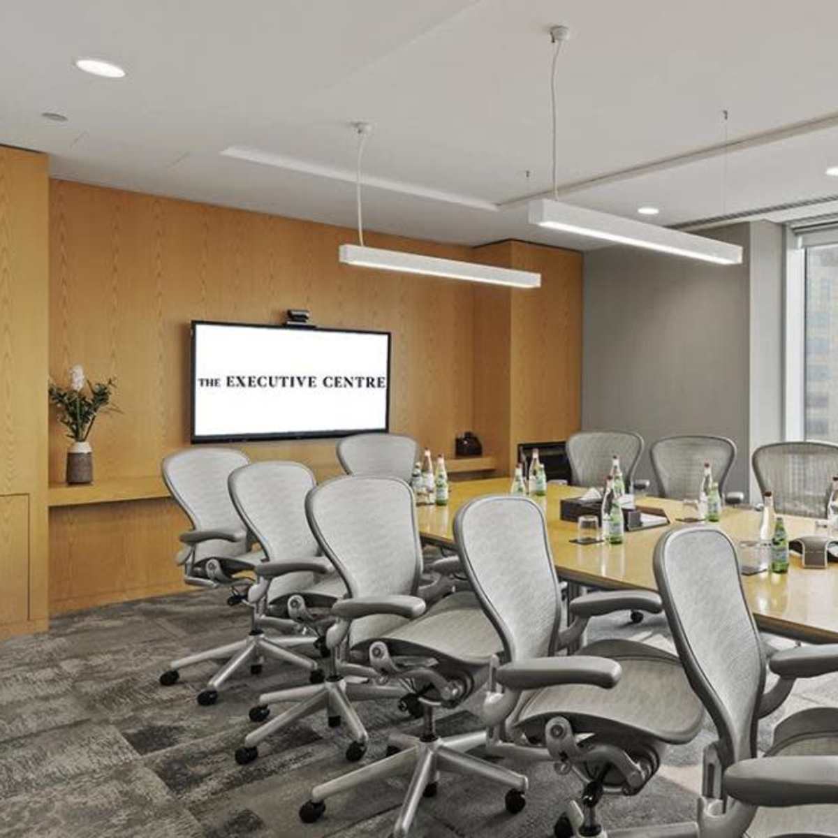 A modern conference room features a long wooden table surrounded by ergonomic chairs, with large windows offering a view of a city skyline.