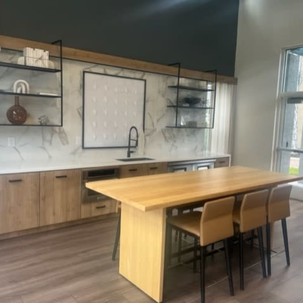 A modern kitchen with wooden cabinets, a large central table, and open shelving displaying decorative items.