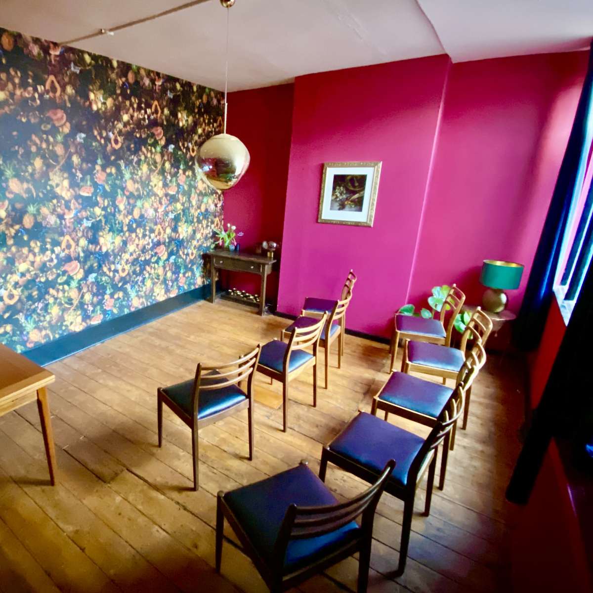 A small room features a floral patterned wall, a wooden table with a glassware set, and several wooden chairs arranged in a semi-circle.