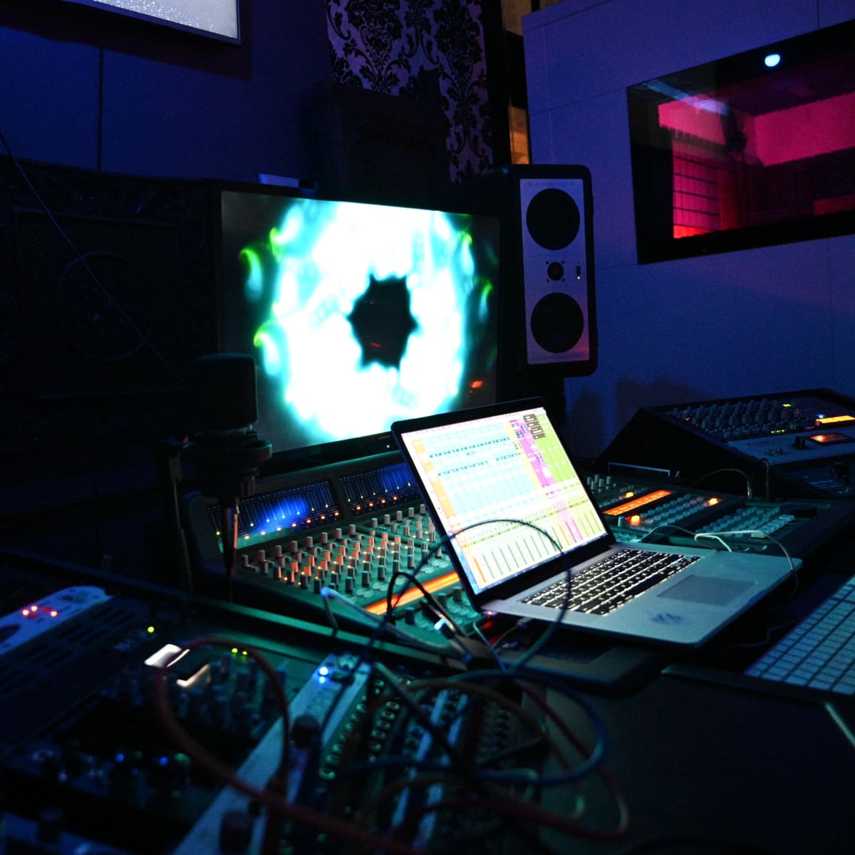 A music production studio features an array of audio equipment, including a mixing console, computer, and large speakers, with a colorful visual display on a screen.