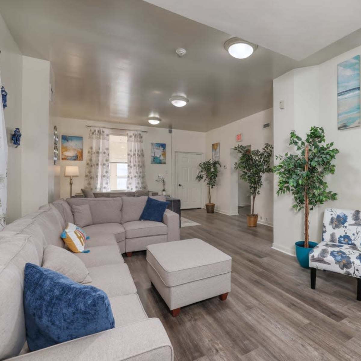 A modern, neutral-toned living room with a sectional sofa, decorative plants, and ocean-themed wall art.