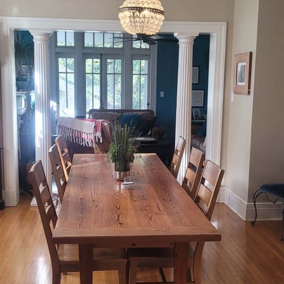 A long wooden dining table with eight chairs is illuminated by a chandelier, set in a brightly lit room featuring large windows and wooden flooring.