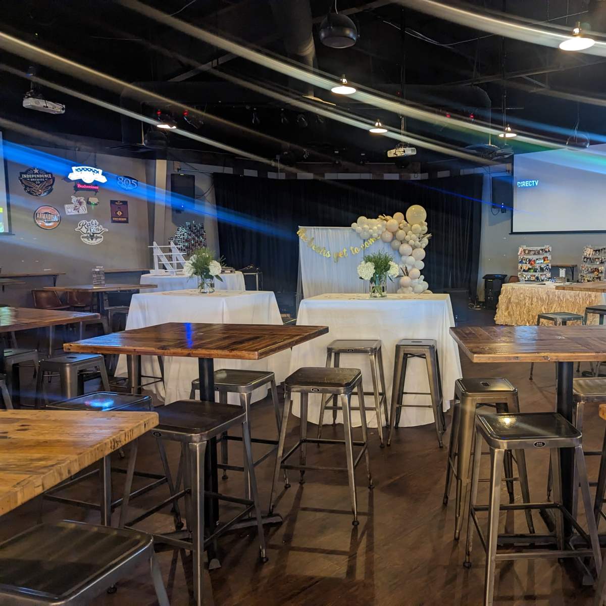 A spacious event venue with several wooden tables and chairs, featuring a decorated stage area and a display table in the background.