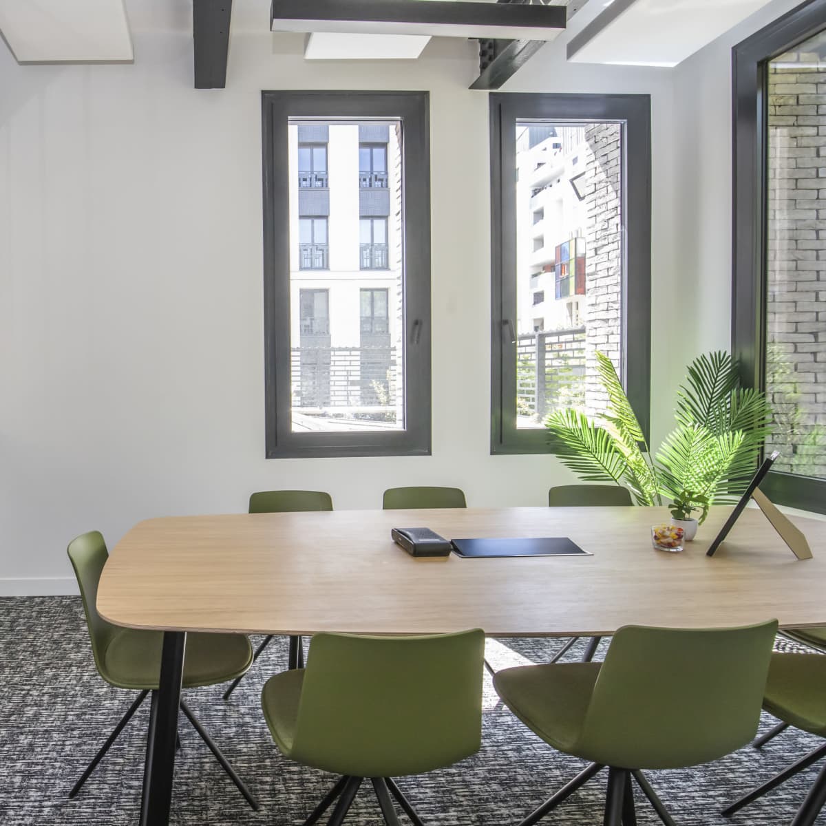 A modern conference room features a wooden table surrounded by green chairs, with large windows allowing natural light to enter.