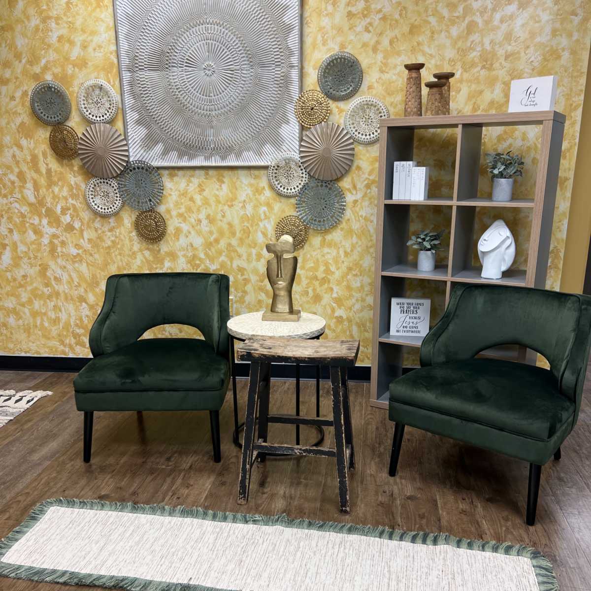 A small seating area with two green chairs, a round table with a decorative sculptural piece, a patterned wall decoration, and a light-colored runner on the wooden floor.