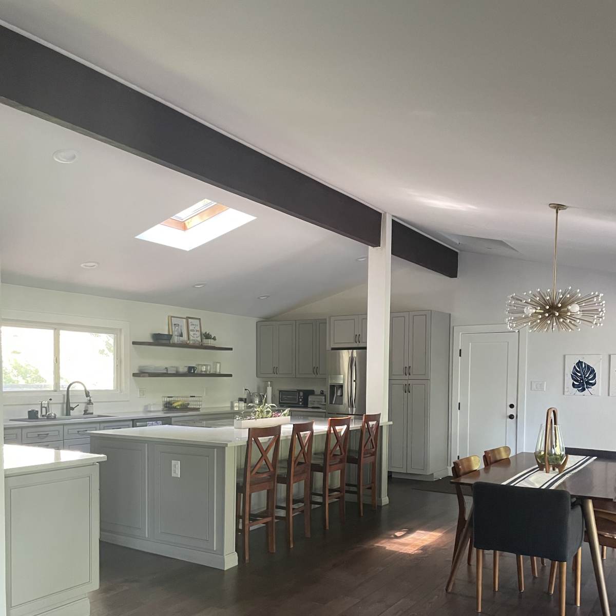 A modern kitchen features a central island with bar stools, light-colored cabinetry, and a dining area with a table and chairs against a backdrop of wooden flooring.