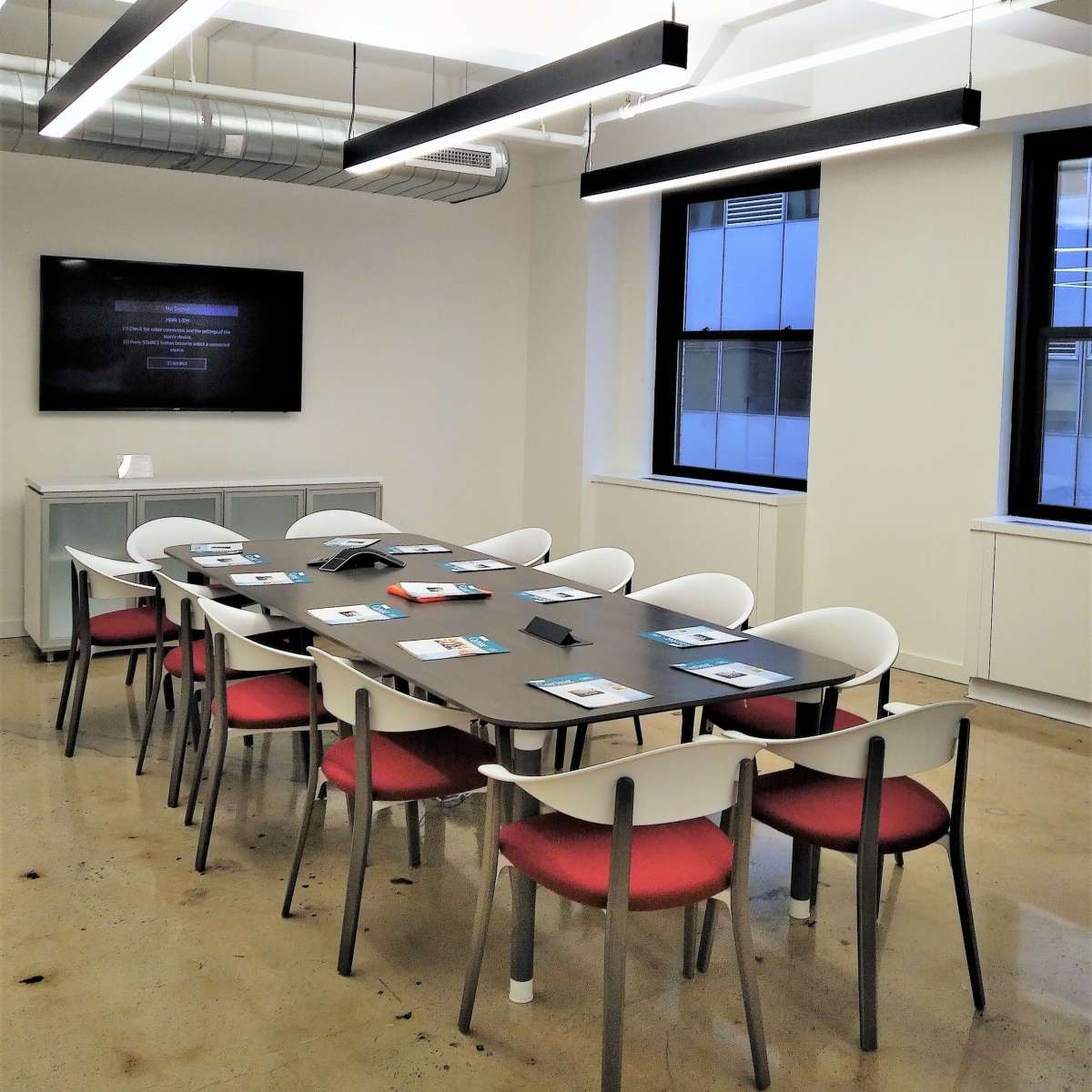 A modern conference room contains a long table surrounded by chairs, with a large screen on one wall and overhead lighting.