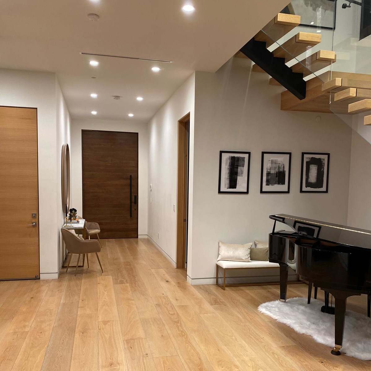 A spacious entryway features light wood flooring, a black grand piano, a console table, and framed artwork along the walls, with stairs leading to an upper level.