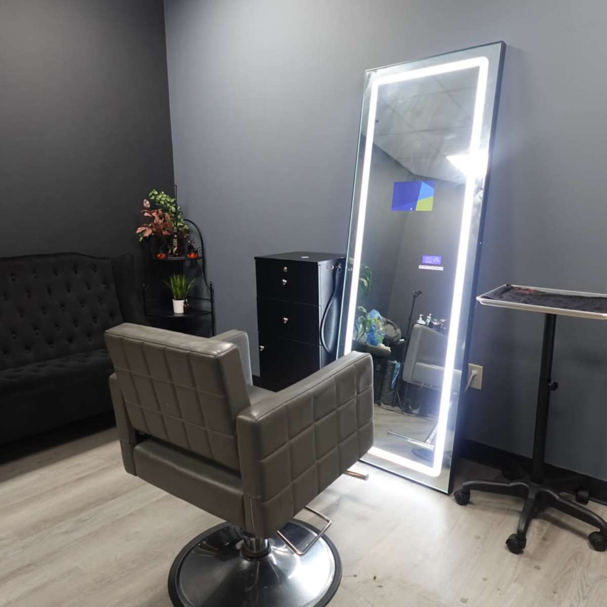 A modern hair salon interior featuring a stylist chair in front of a large illuminated mirror, with a black cabinet and seating in the background.