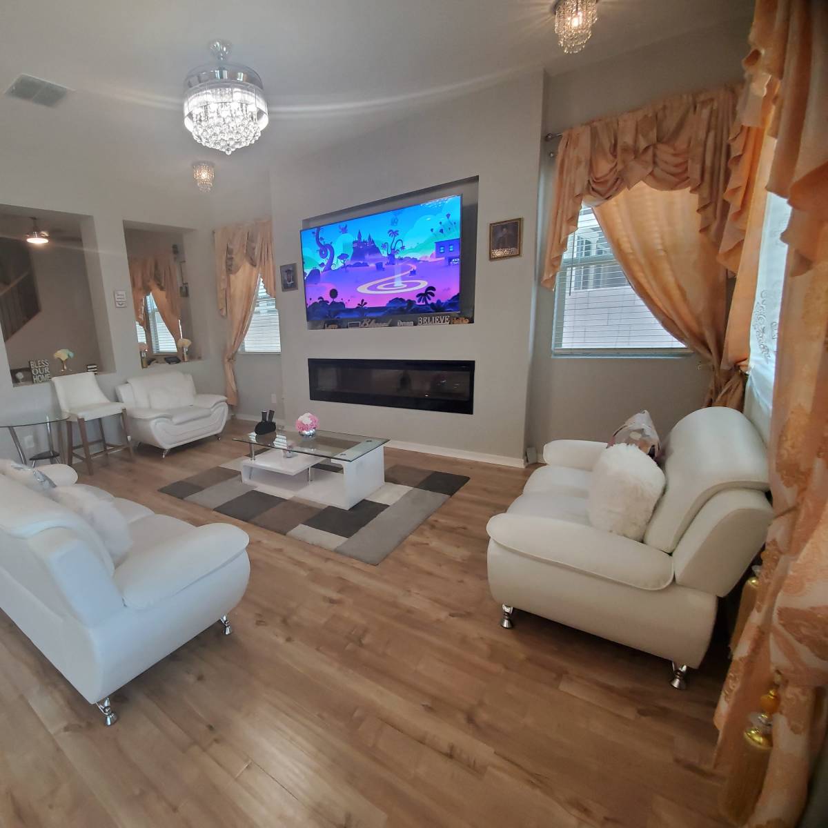 The living room features two white sofas, a glass coffee table, and a large wall-mounted TV, with decorative curtains and a modern fireplace.