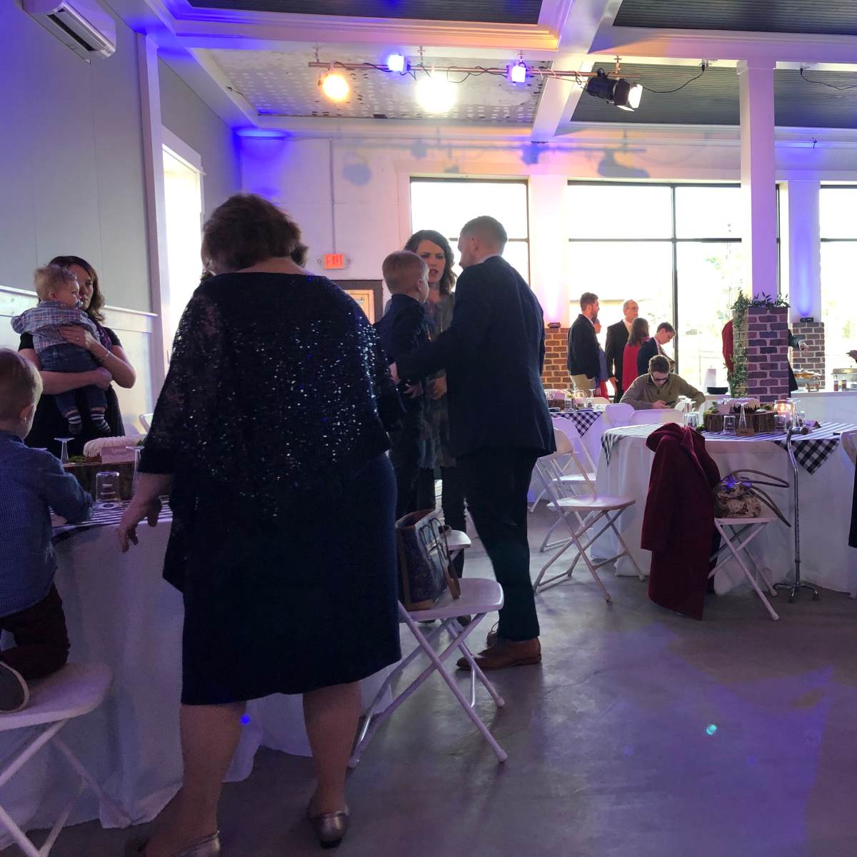 A spacious event venue with guests interacting and seated at white tables, while a few people stand around, surrounded by decorations and a large window letting in natural light.