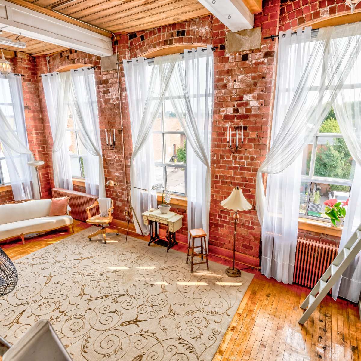 A spacious room with large windows dressed in sheer curtains, brick walls, wooden floors, and a mix of furniture including a sofa, chairs, and a lamp.