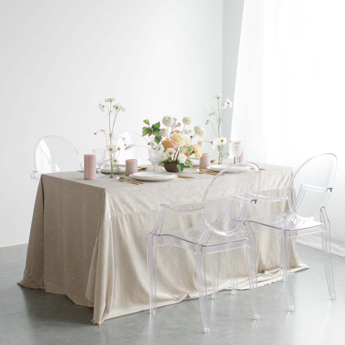A dining table set with a neutral tablecloth, plates, and vases of flowers, surrounded by transparent chairs.