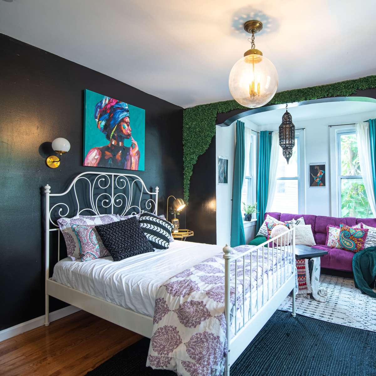 A stylish bedroom featuring a white metal bed, dark walls, vibrant artwork, and a cozy seating area with plants and decorative textiles.