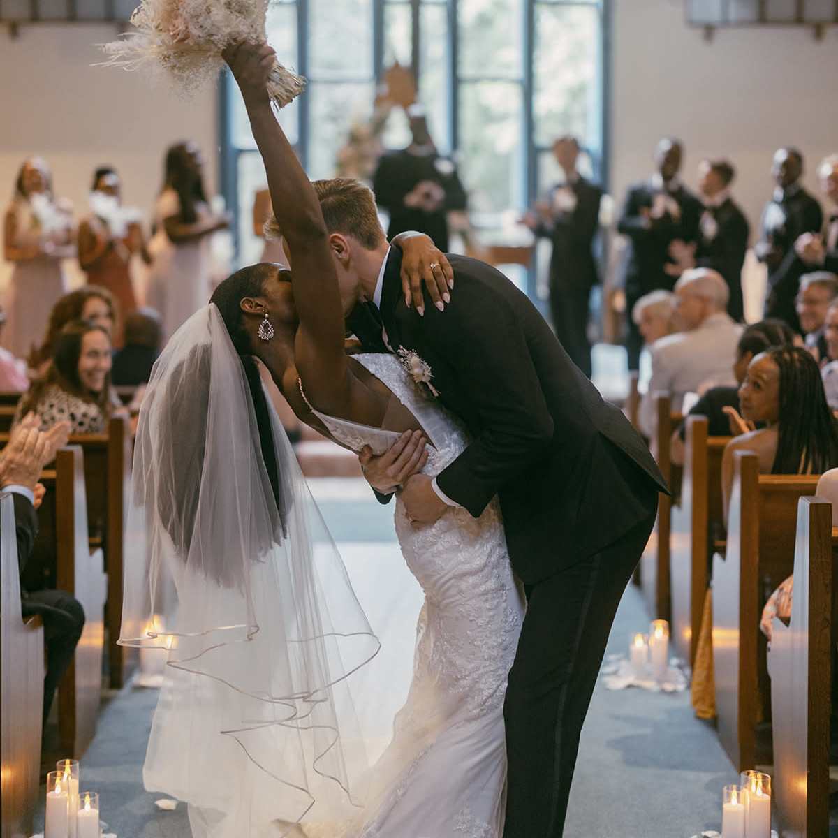 A bride and groom kiss while she raises her bouquet, surrounded by seated guests in a church.