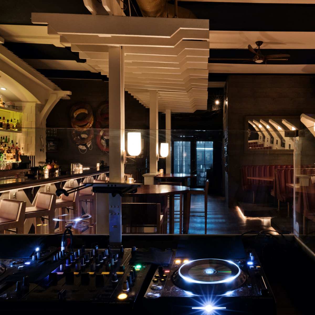 A modern bar and restaurant interior featuring a DJ setup in the foreground with illuminated tables and booths in a dimly lit atmosphere.