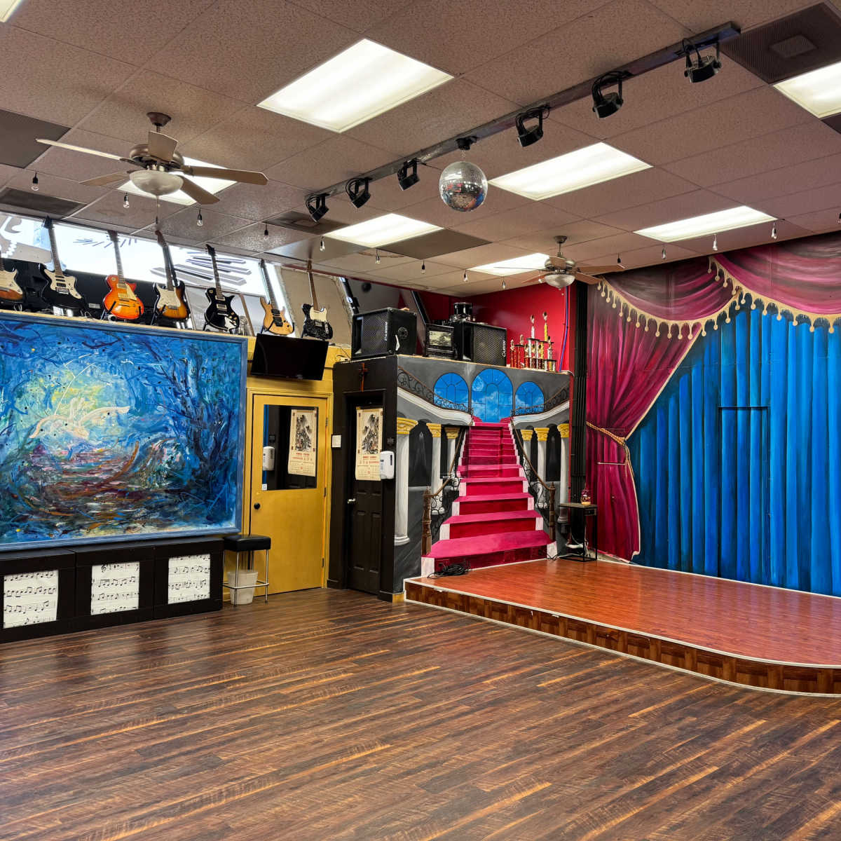 A performance space with a wooden stage, a painted blue curtain backdrop, a staircase, and musical instruments displayed on the walls.