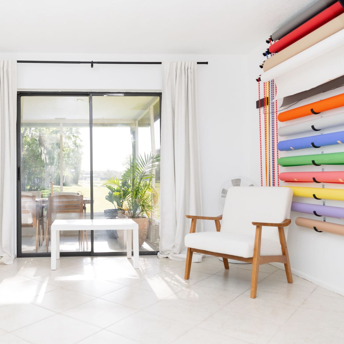 A bright, minimalist room features a chair and a small table near a glass sliding door overlooking a patio and greenery.
