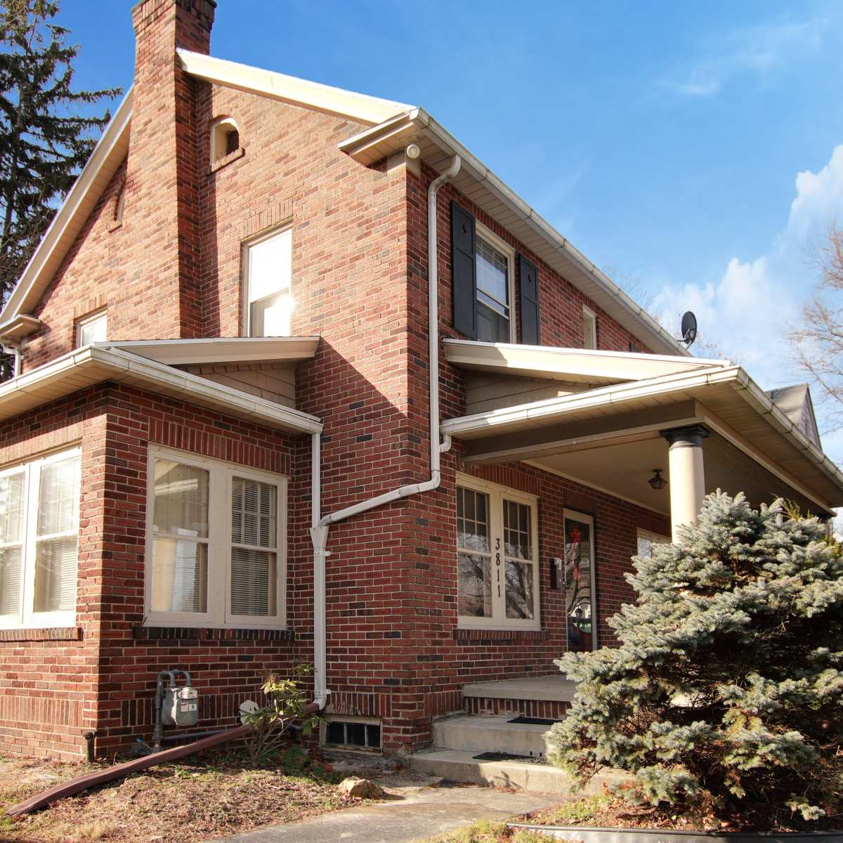 A two-story brick house features a front porch and large windows, surrounded by trees and a landscaped yard.