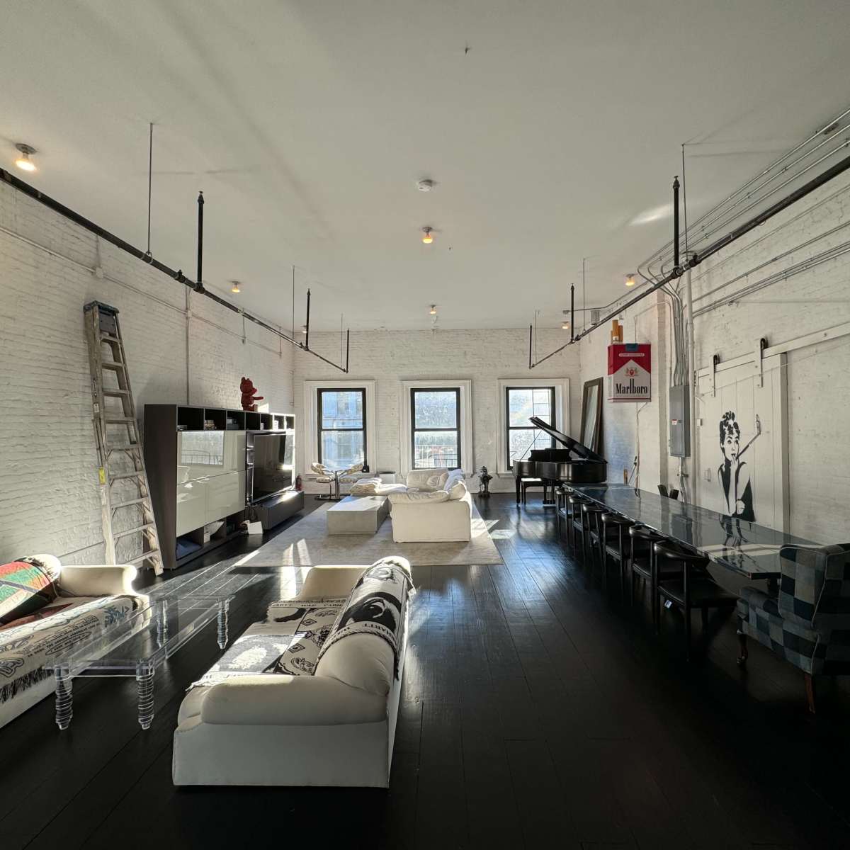 A spacious, modern loft featuring white brick walls, a long dining table, clear acrylic furniture, and a grand piano, with large windows allowing natural light to fill the space.