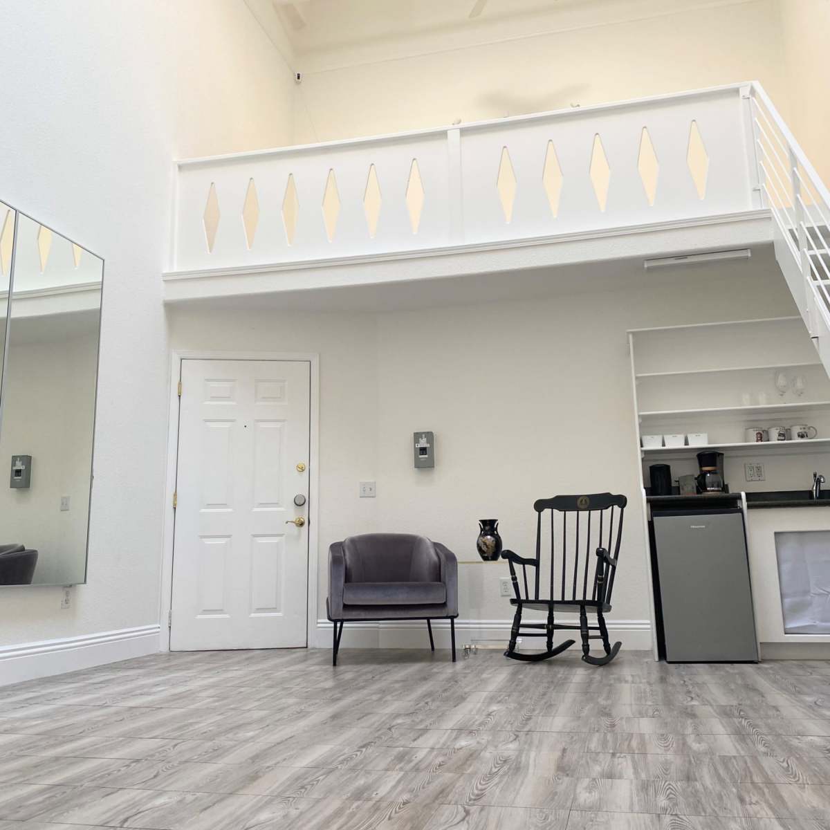 A spacious room with a high ceiling, a rocking chair, a small refrigerator, and a door leading outside, along with a mirrored wall and a mezzanine balcony.