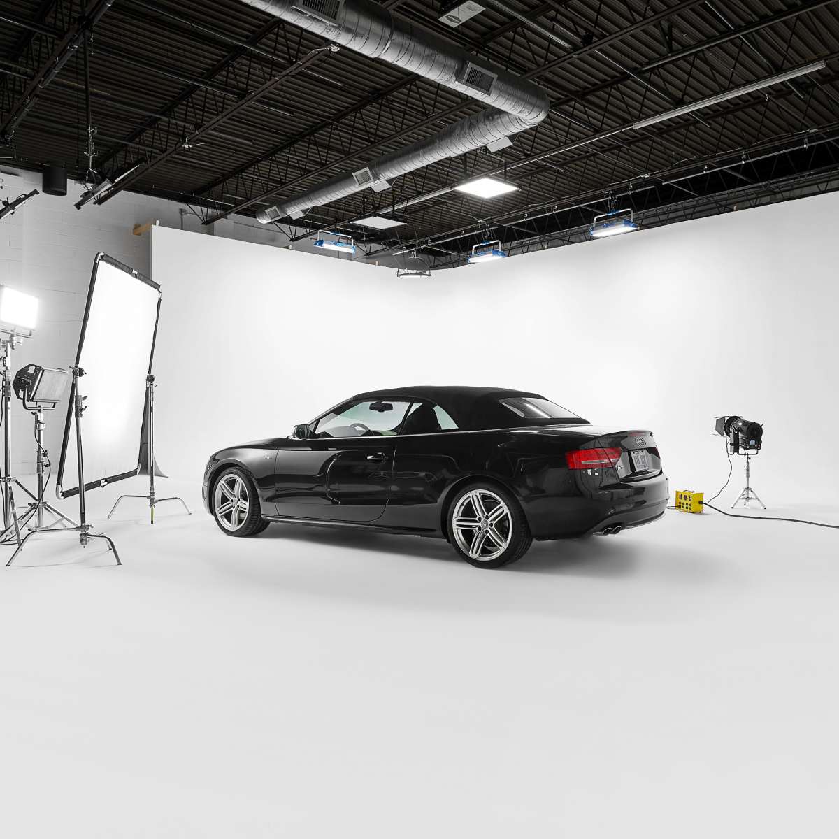 A black car is positioned in a studio with a white backdrop and professional lighting equipment.