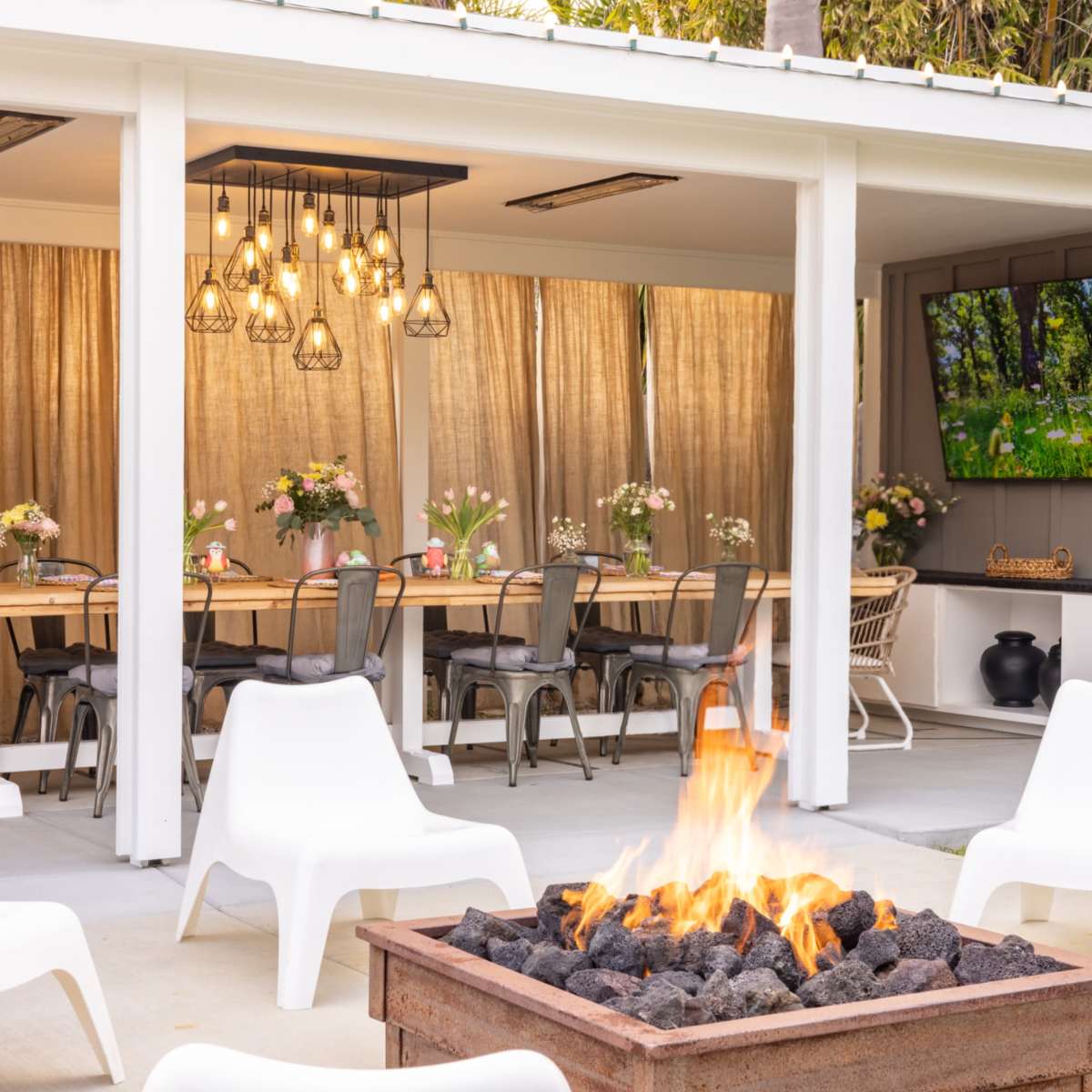 An outdoor seating area features a fire pit at the center, surrounded by white chairs, with a long dining table set beneath warm-toned pendant lights and decorative curtains.